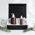 Two skincare products in a black box with white tissue paper, surrounded by cotton and eucalyptus leaves on a light background.